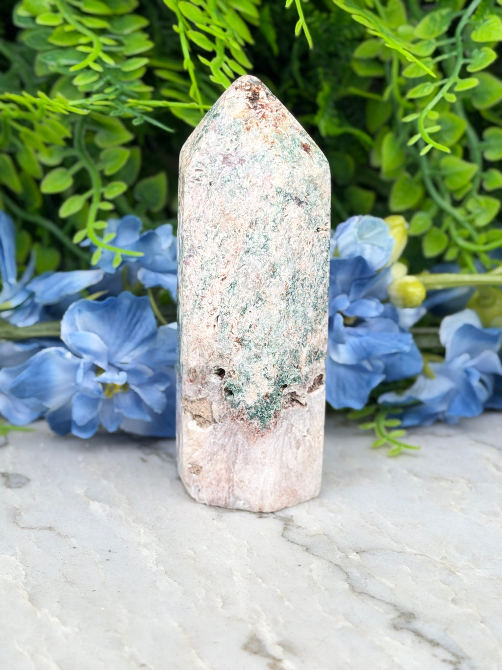 A white, moss-covered stone sits on a marble surface, surrounded by vibrant blue flowers.