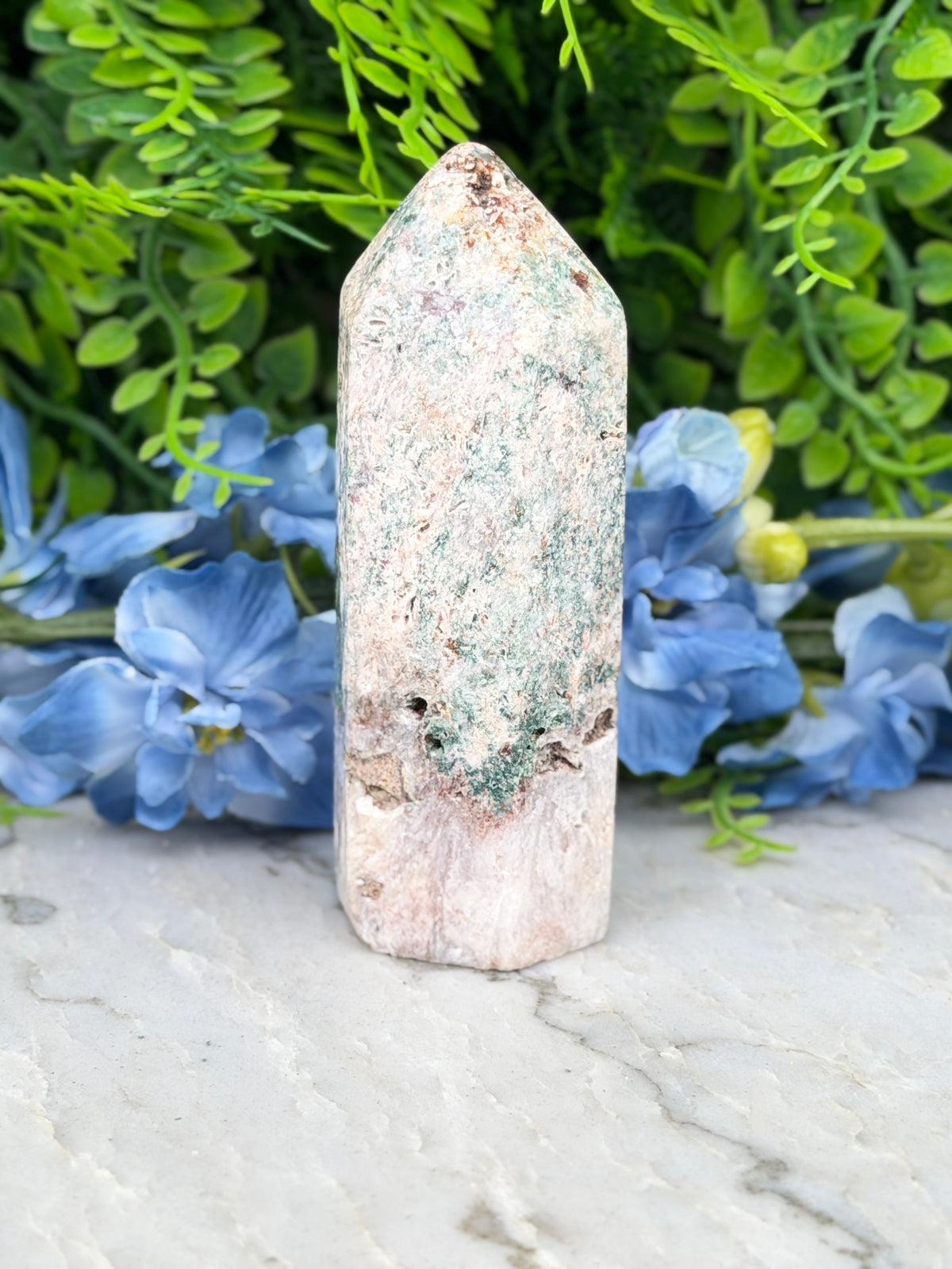 A white, moss-covered stone sits on a marble surface, surrounded by vibrant blue flowers.