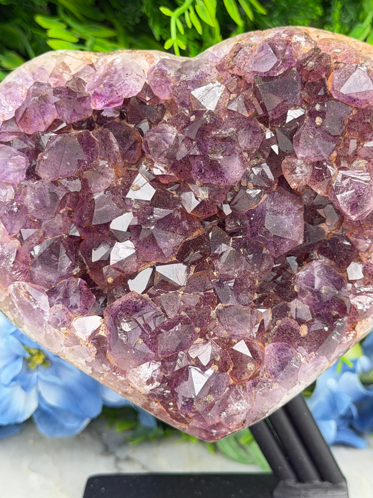 a close-up view of a large, clear crystal with numerous small, sharp-edged crystals embedded within it. The crystal appears to be purple in color, and it is set against a backdrop of green foliage and blue flowers.