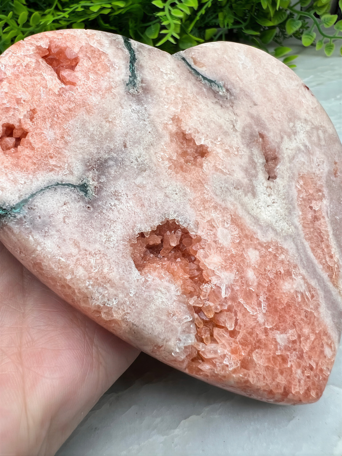 A hand is holding a heart-shaped piece of pink crystal, which appears to be a natural formation.