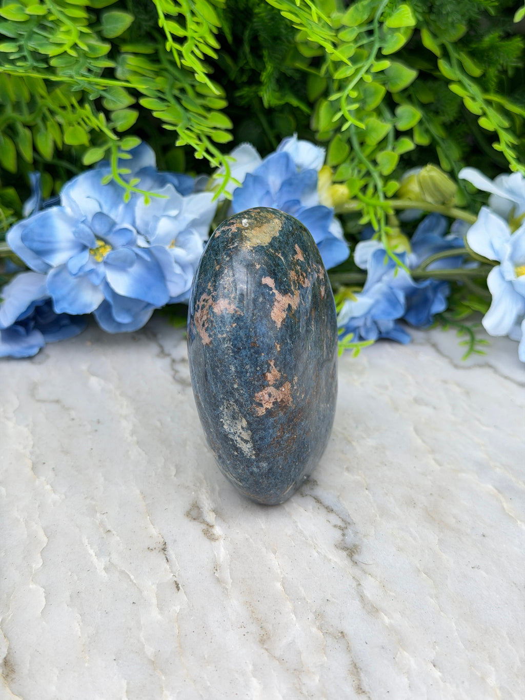 a close-up of a blue and black rock resting on a white surface, surrounded by lush green foliage and delicate blue flowers.