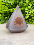 A gray, triangular crystal with a brownish-brown stone in the center sits on a white surface, surrounded by lush green foliage.