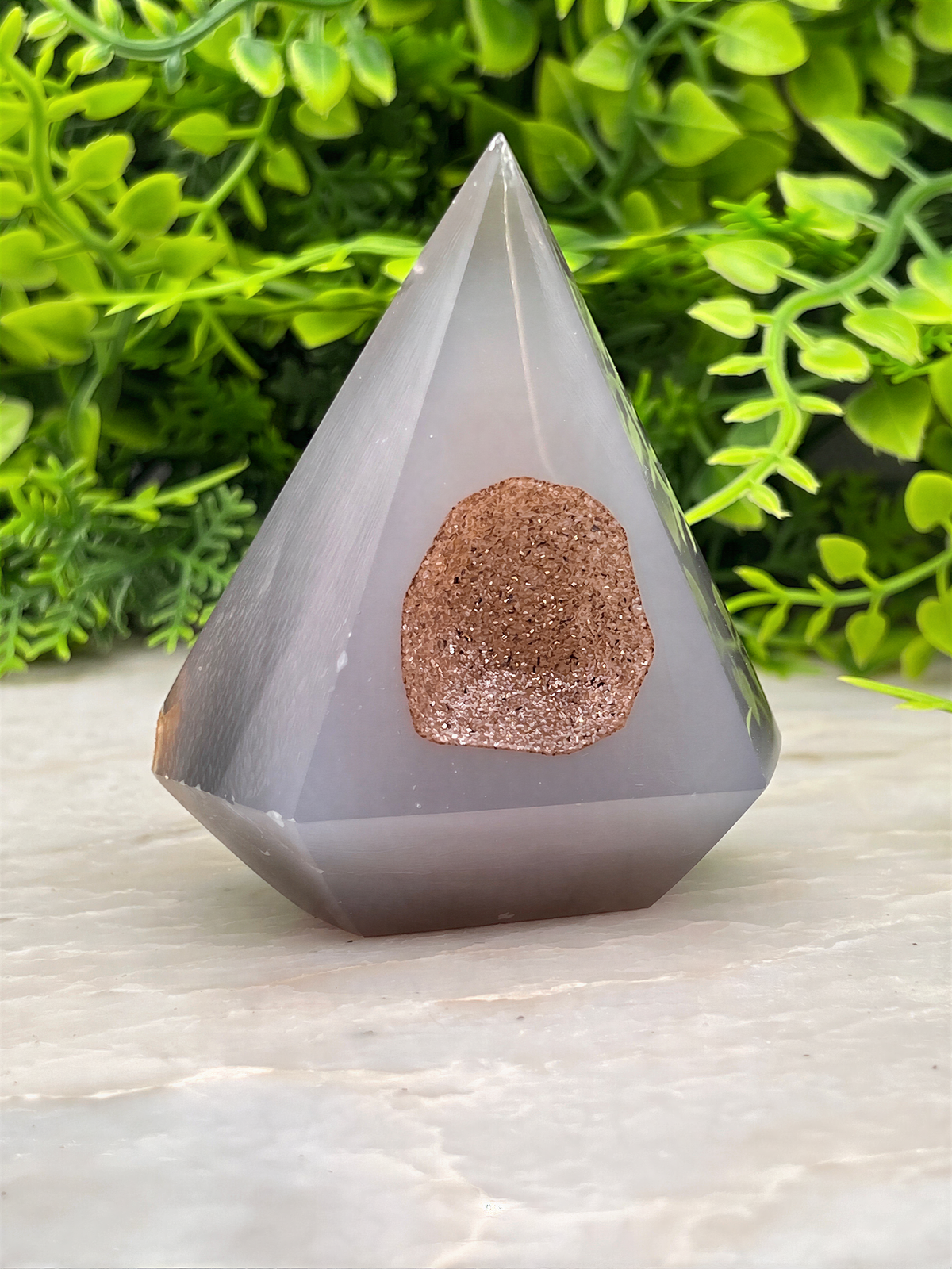 A gray, triangular crystal with a brownish-brown stone in the center sits on a white surface, surrounded by lush green foliage.