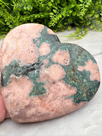 A heart-shaped rock with a mix of green and pink hues, with a textured surface, is displayed against a backdrop of greenery.
