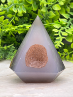A gray, triangular crystal with a brown, irregularly shaped piece of rock embedded in it, set against a backdrop of green foliage.