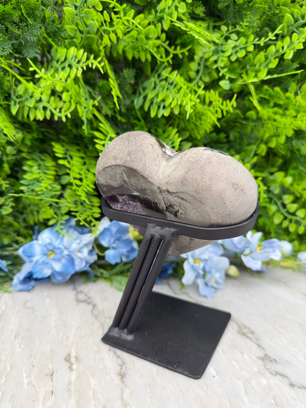 A heart-shaped stone is mounted on a black metal bracket, surrounded by lush green foliage and blue flowers.