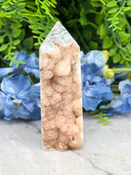 A tall, pointed crystal formation with a rough, textured surface sits in the center of the image, surrounded by a backdrop of lush green foliage and delicate blue flowers.