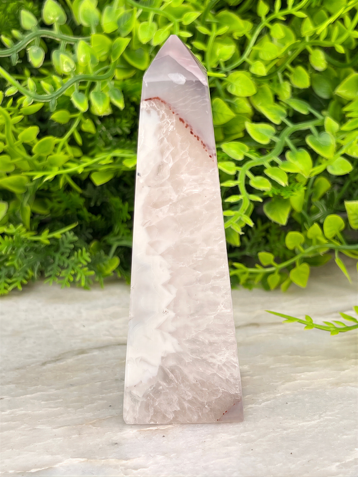 A pink quartz crystal stands upright on a white surface, surrounded by lush green foliage.