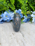 a close-up view of a dark blue rock with a speckled surface, resting on a white surface. The rock is surrounded by a variety of green and blue flowers, creating a visually appealing contrast between the natural and man-made elements.