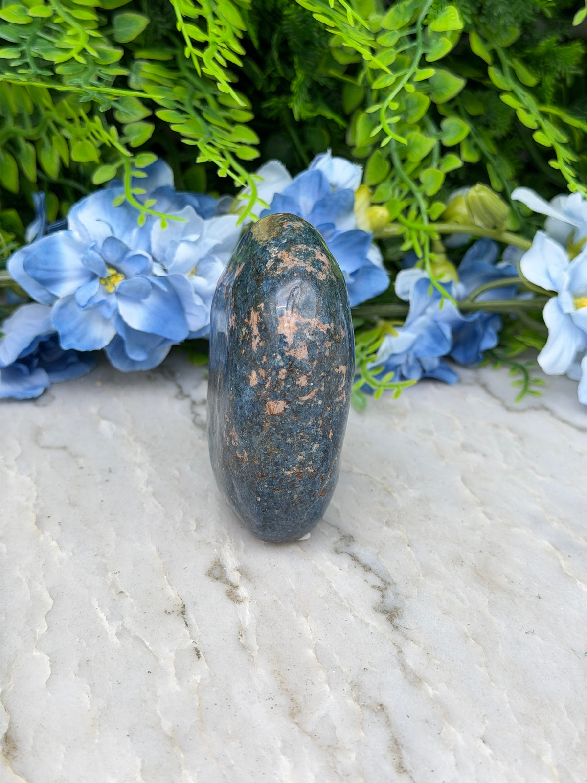 a close-up view of a dark blue rock with a speckled surface, resting on a white surface. The rock is surrounded by a variety of green and blue flowers, creating a visually appealing contrast between the natural and man-made elements.