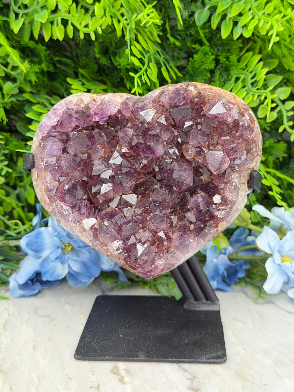 A heart-shaped crystal formation with numerous small purple crystals is displayed against a backdrop of green foliage and blue flowers.