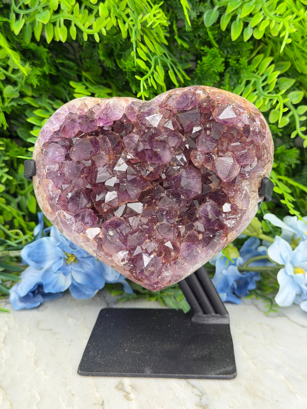A heart-shaped crystal formation with numerous small purple crystals is displayed against a backdrop of green foliage and blue flowers.