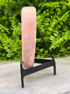 A pink crystal or gemstone is displayed on a black metal stand, set against a backdrop of green foliage.