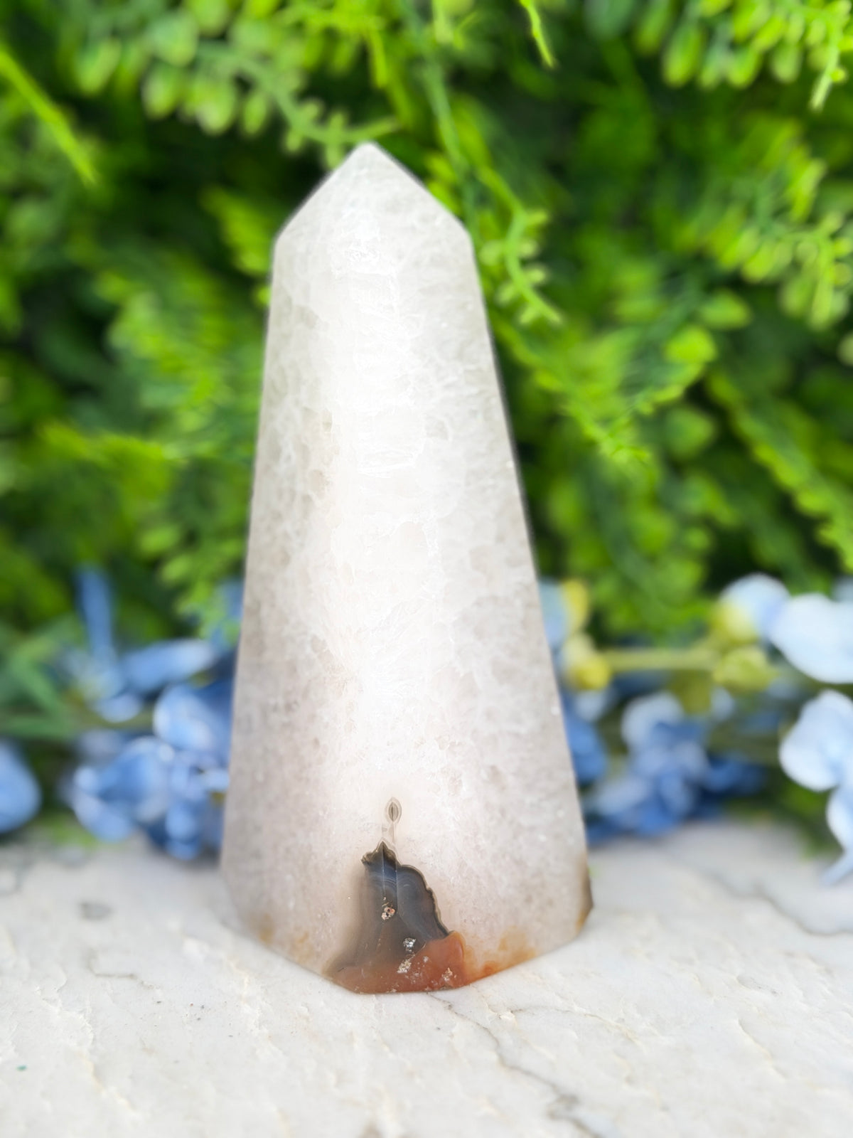A white crystal formation with a small hole in the center, surrounded by a blurred green background with blue flowers.