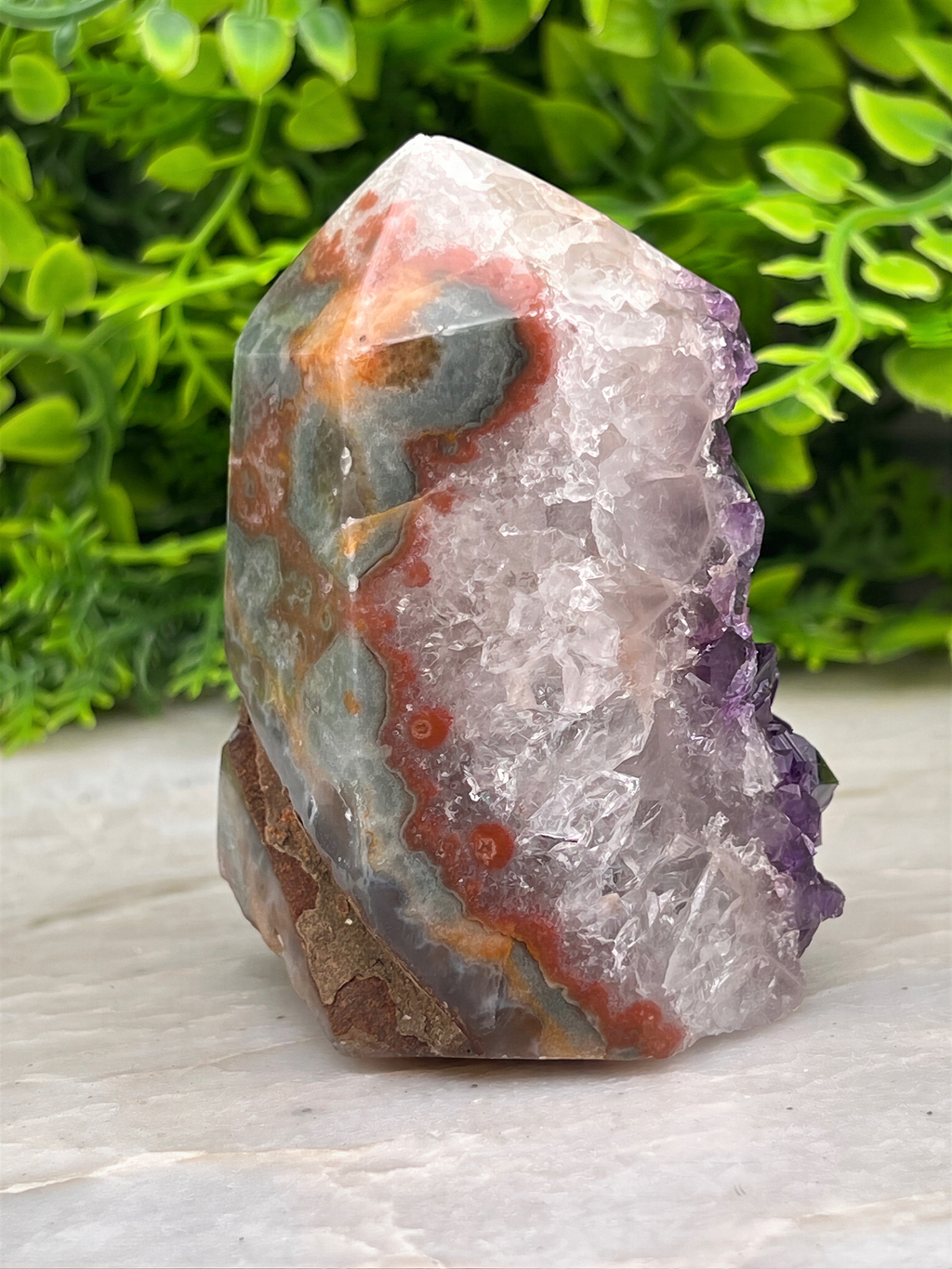 a large, colorful crystal formation with a mix of white, orange, and purple hues, set against a backdrop of green foliage.