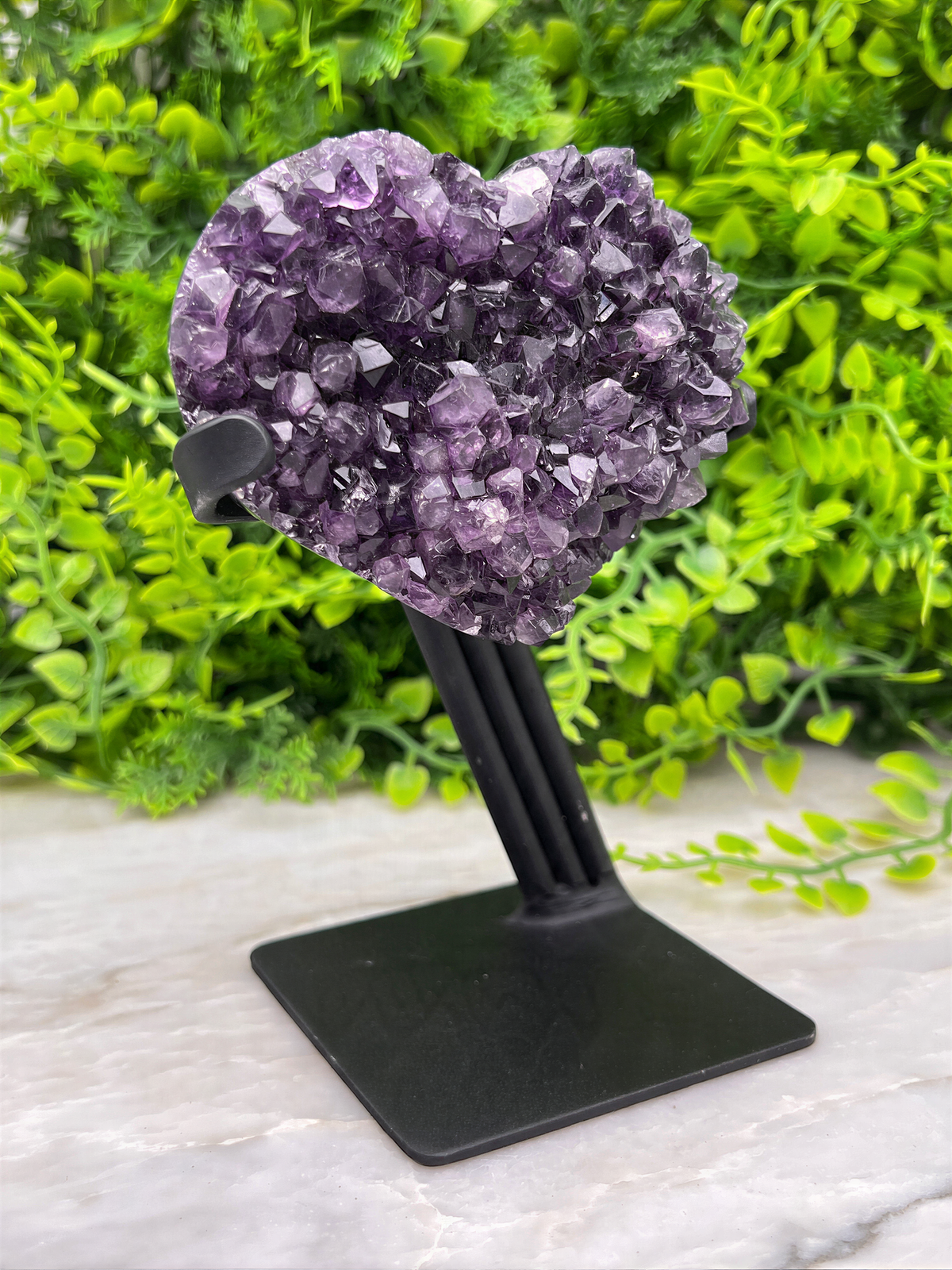 A heart-shaped cluster of purple amethyst crystals is displayed on a black stand, surrounded by lush green foliage.