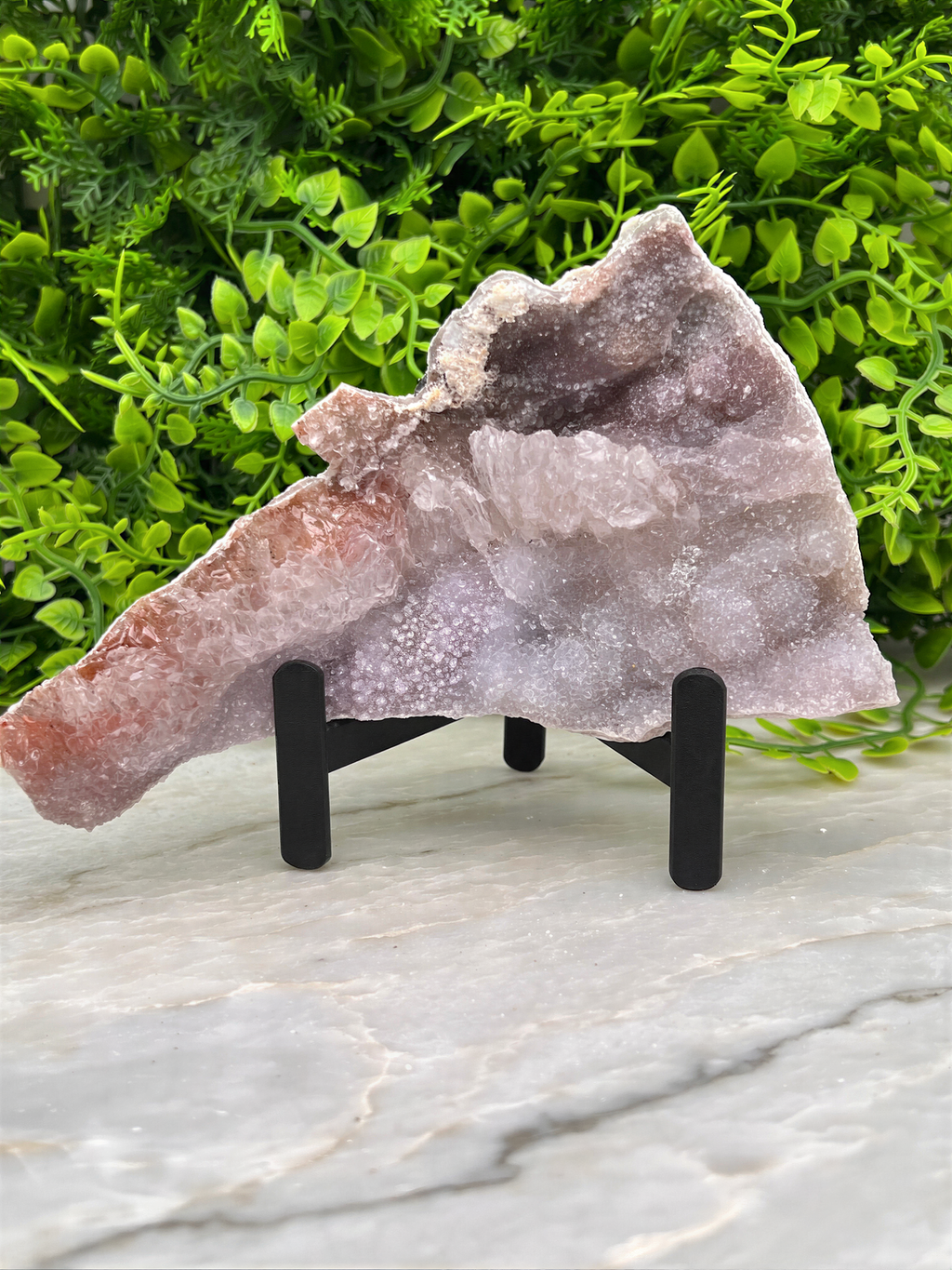 a large piece of amethyst, a purple crystal, displayed on a black stand in front of a lush green plant.