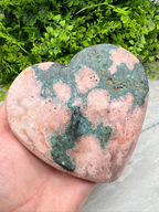 A hand is holding a heart-shaped rock with a colorful, speckled surface, set against a backdrop of lush green foliage.