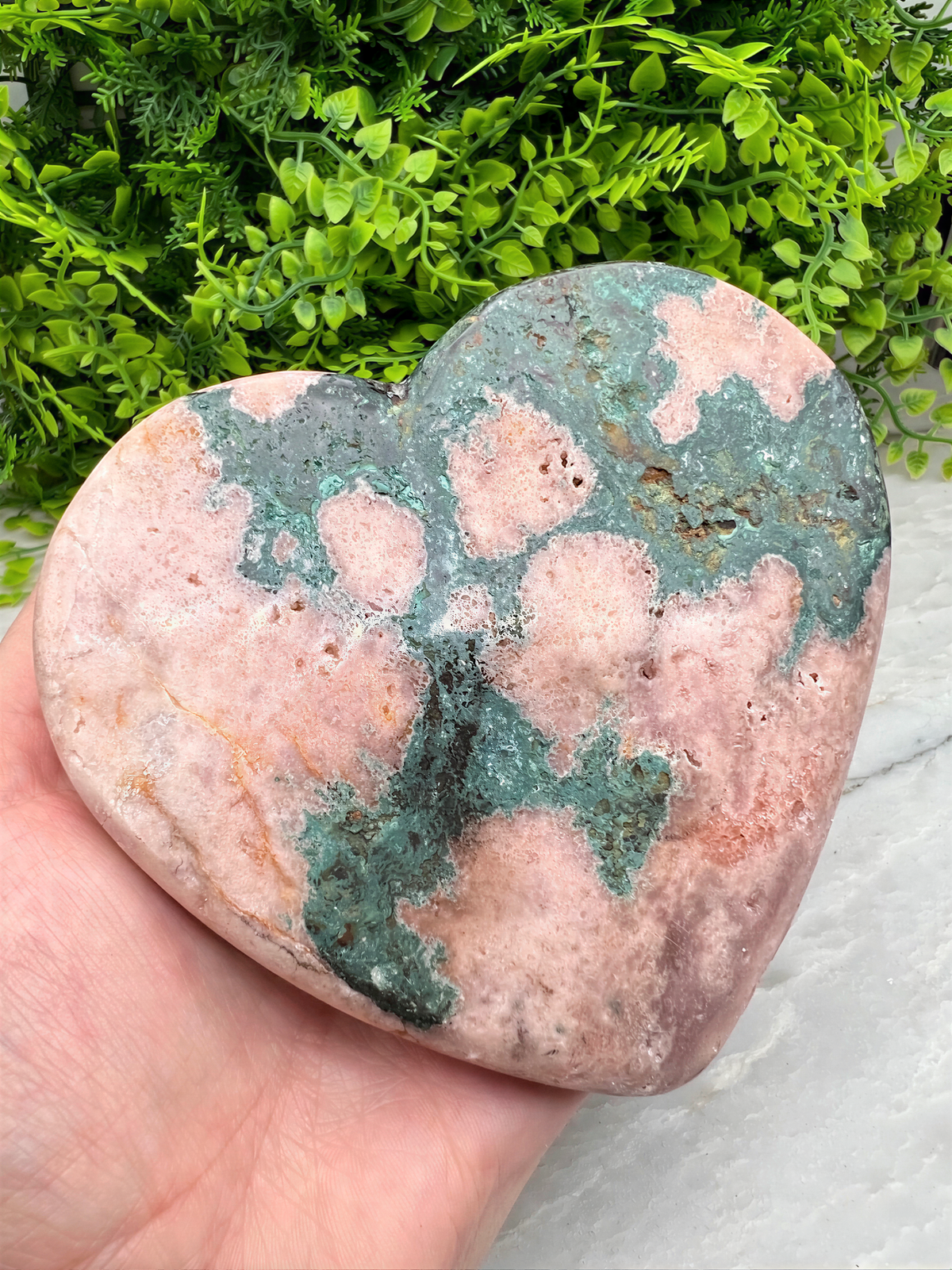 A hand is holding a heart-shaped rock with a colorful, speckled surface, set against a backdrop of lush green foliage.