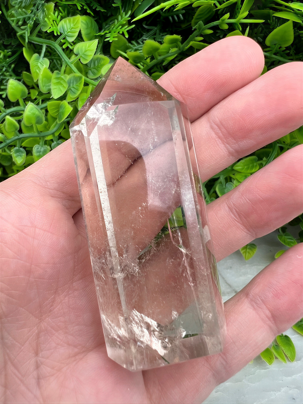 A hand holding a clear crystal with a jagged edge, set against a background of green foliage.