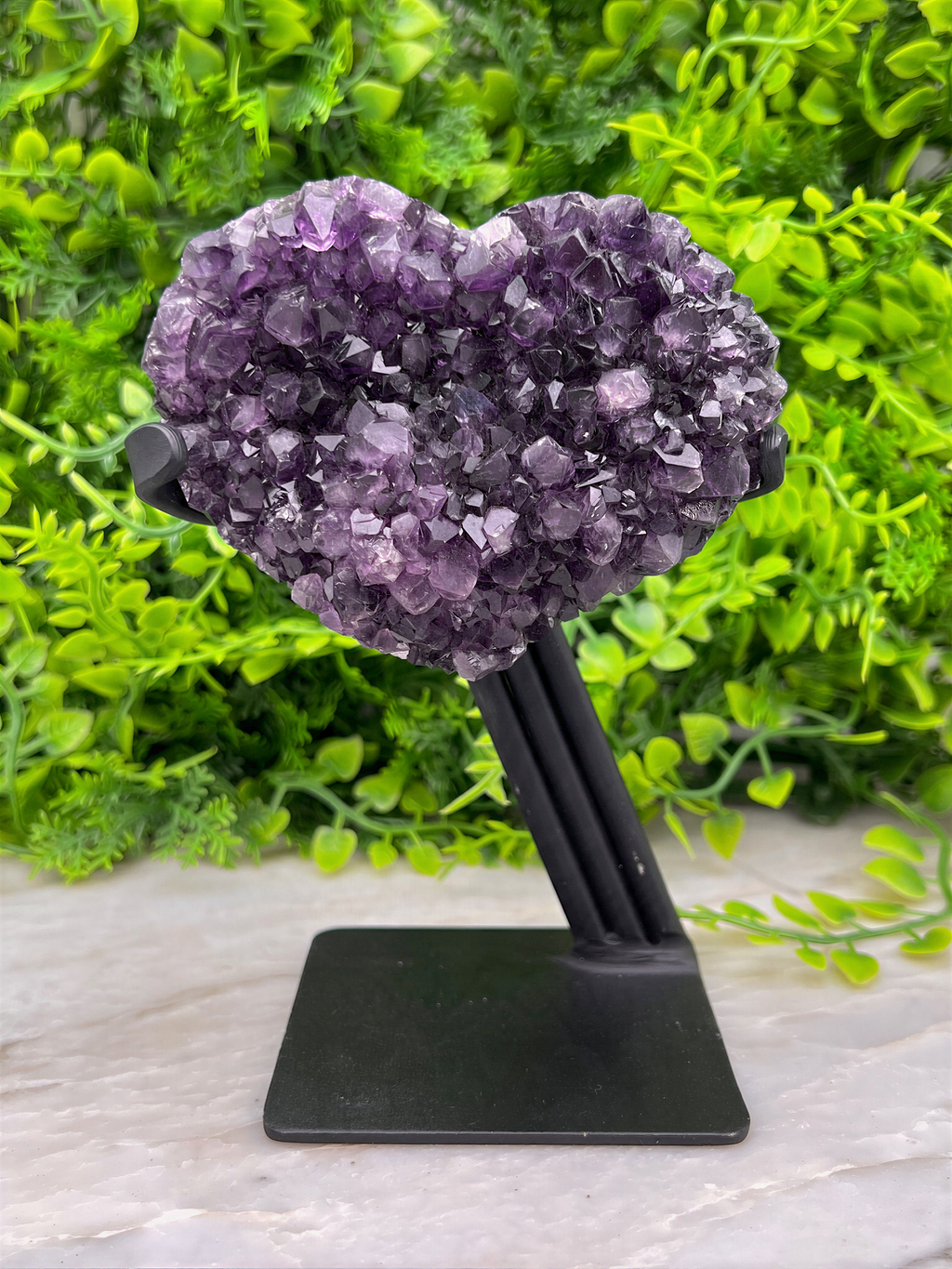 A heart-shaped cluster of purple amethyst crystals is displayed on a black stand, set against a backdrop of lush green foliage.