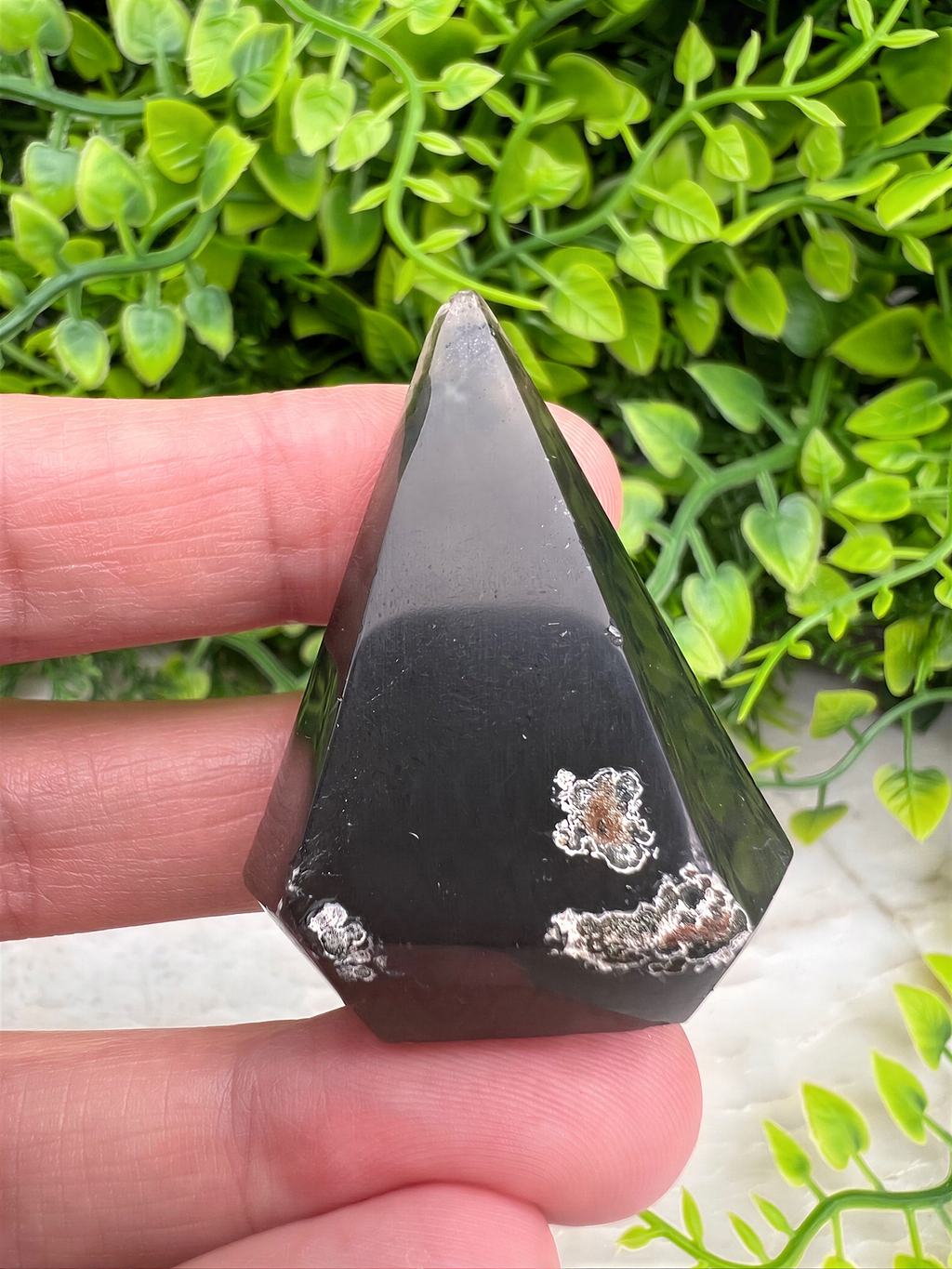 A hand holding a black, triangular-shaped crystal against a background of green foliage.