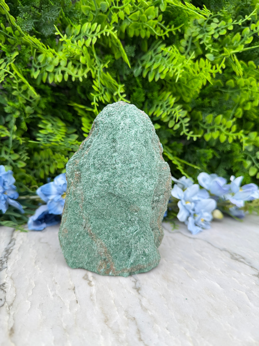 A large, green rock lies on a white surface, surrounded by lush green foliage and delicate blue flowers.