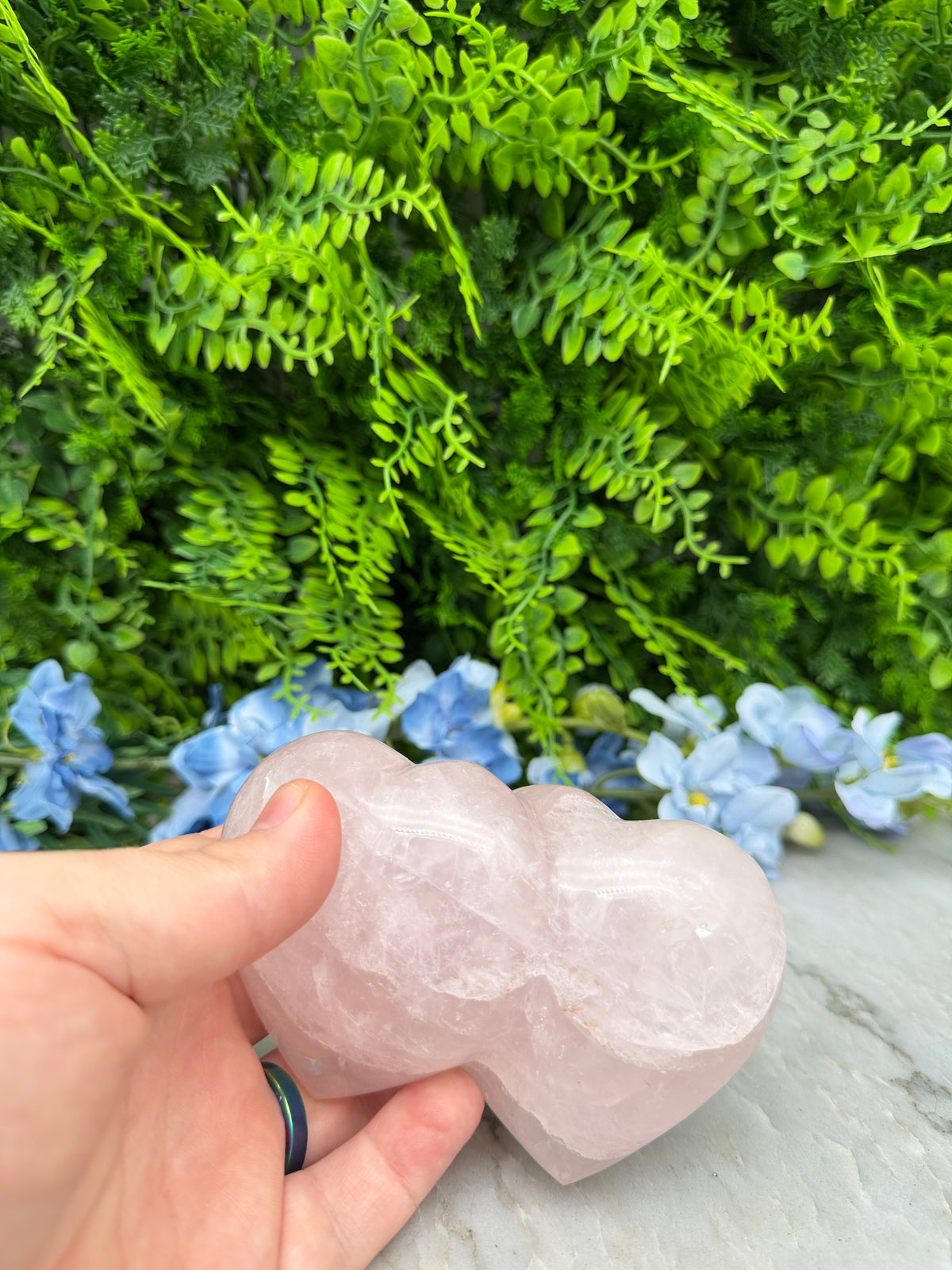 A hand holding a pink crystal against a backdrop of green foliage and blue flowers.