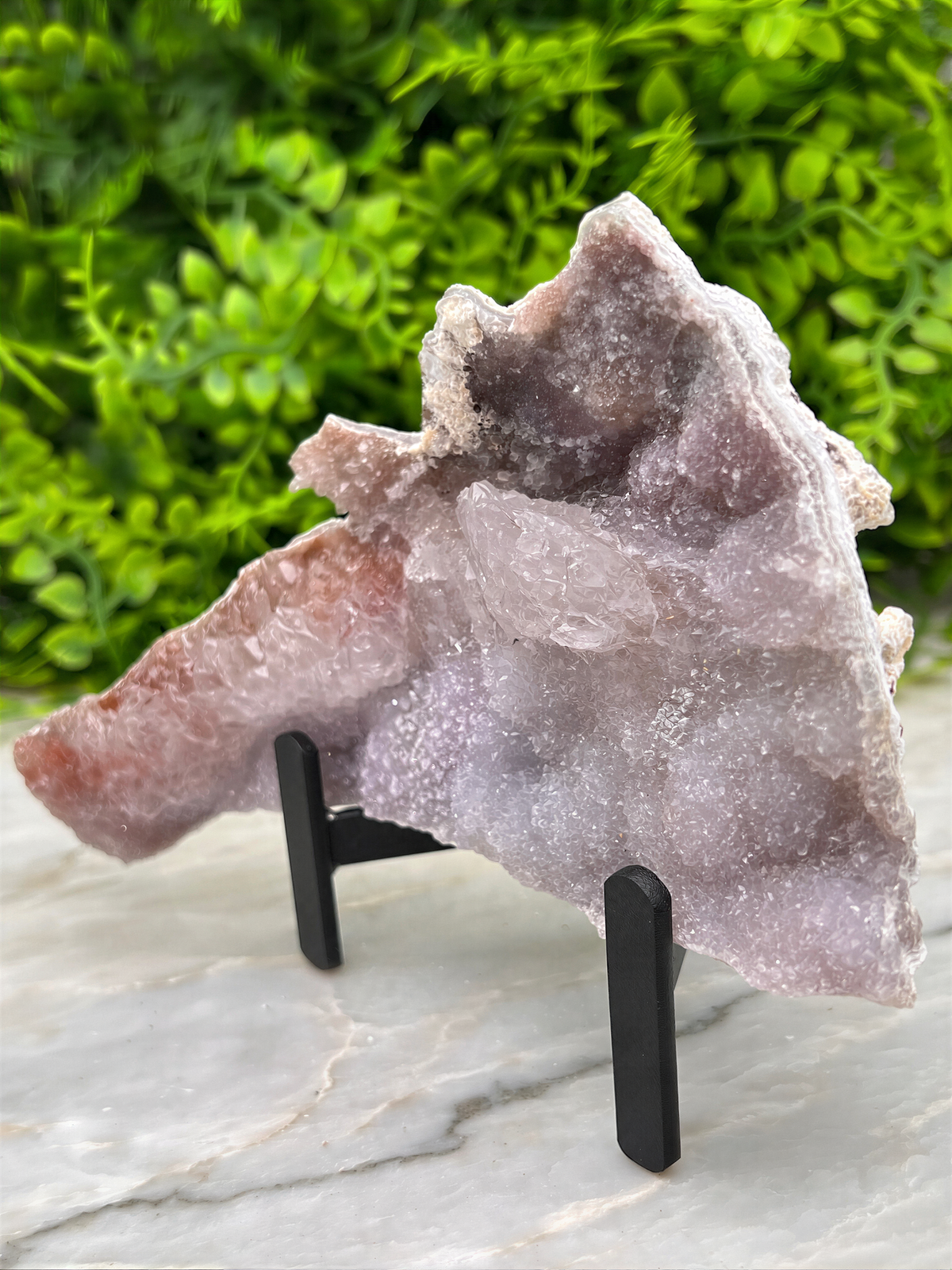 a large piece of amethyst, a purple crystal, displayed on a stand. The crystal is set against a backdrop of green foliage, creating a visually appealing contrast.