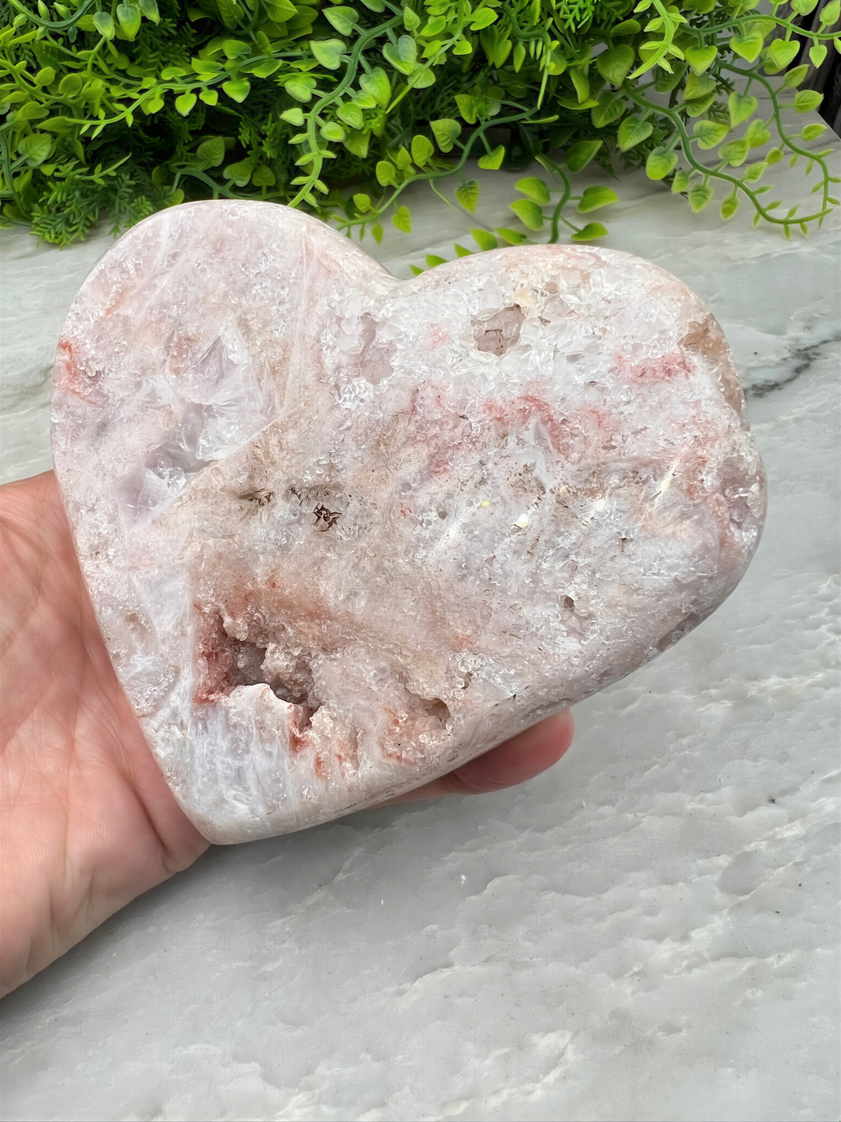 A hand is holding a heart-shaped rock with a pinkish-white color and a rough, jagged texture. The rock is placed on a white surface, with a green plant visible in the background.
