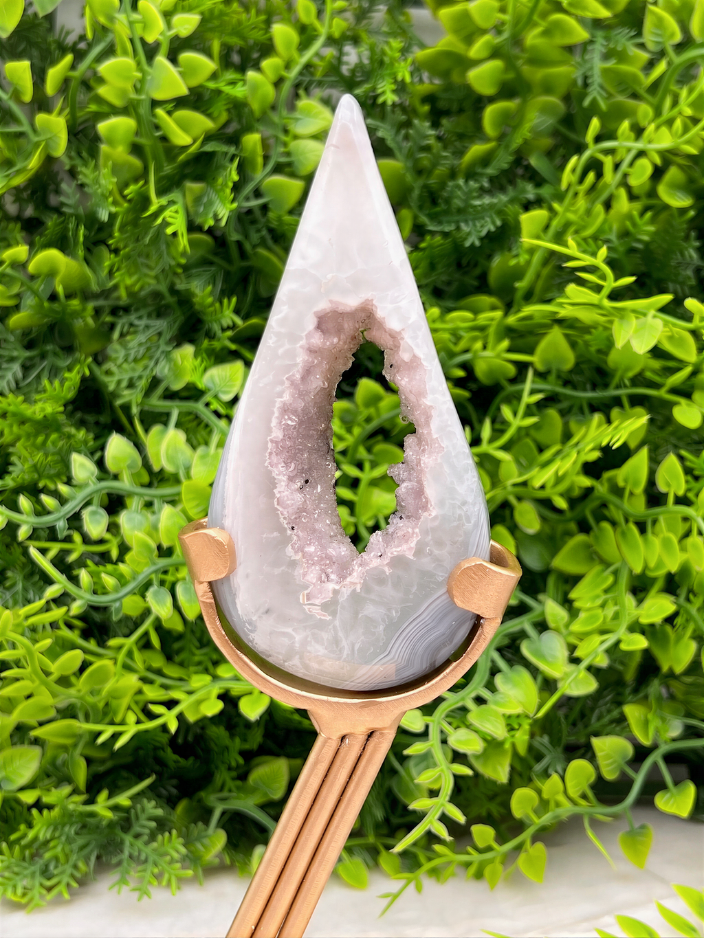 A white geode with a pinkish-purple interior is displayed on a gold stand, surrounded by lush green foliage.