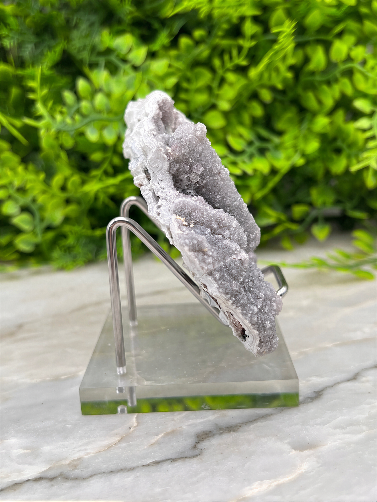 a clear glass stand holding a piece of white crystal, which appears to be a type of quartz. The crystal is placed on a marble surface, and there are some greenery visible in the background.