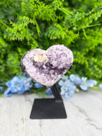 A heart-shaped amethyst crystal on display, surrounded by blue flowers and a green plant.