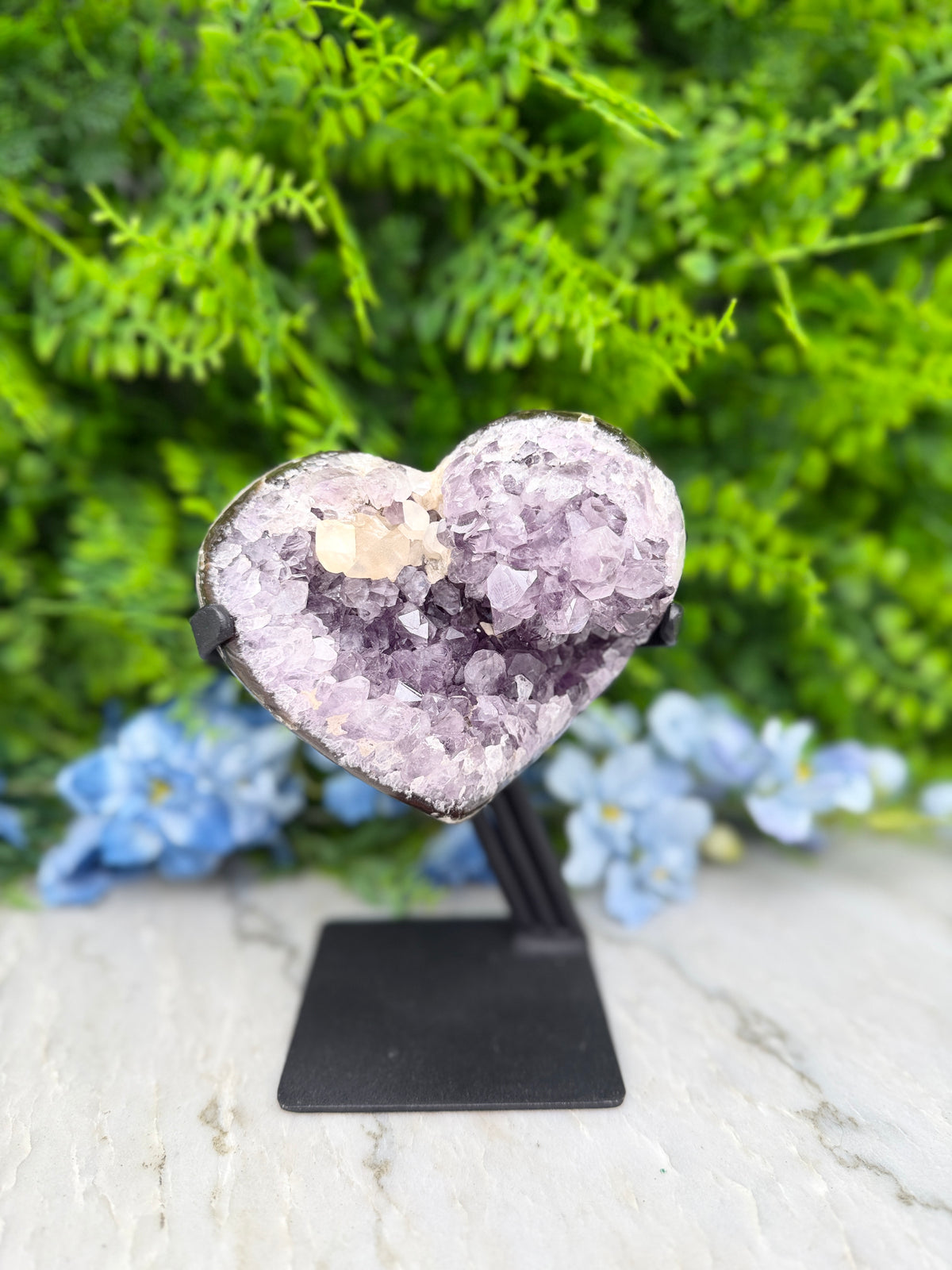 A heart-shaped amethyst crystal on display, surrounded by blue flowers and a green plant.