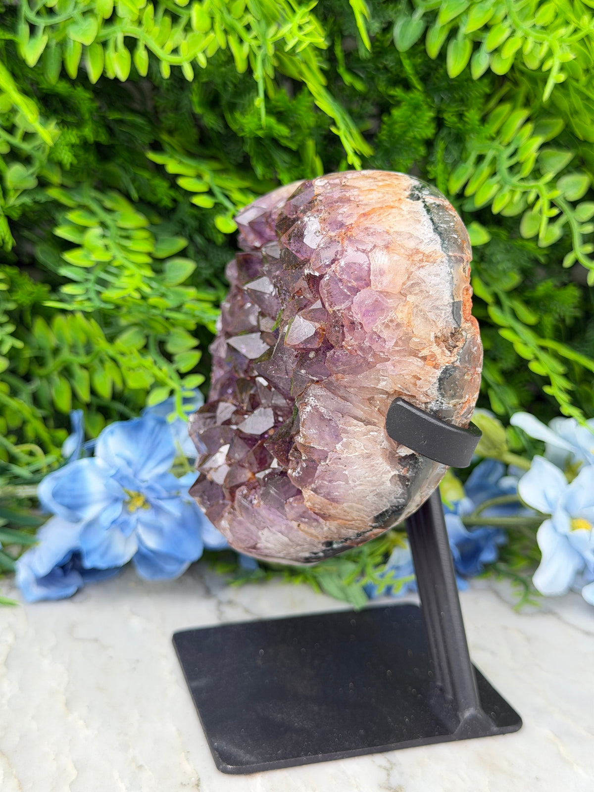 A large, multicolored crystal formation is displayed on a black stand, surrounded by lush green foliage and blue flowers.