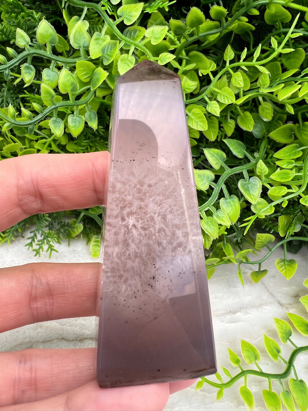 A hand is holding a clear, rectangular-shaped crystal or gemstone against a backdrop of green foliage.