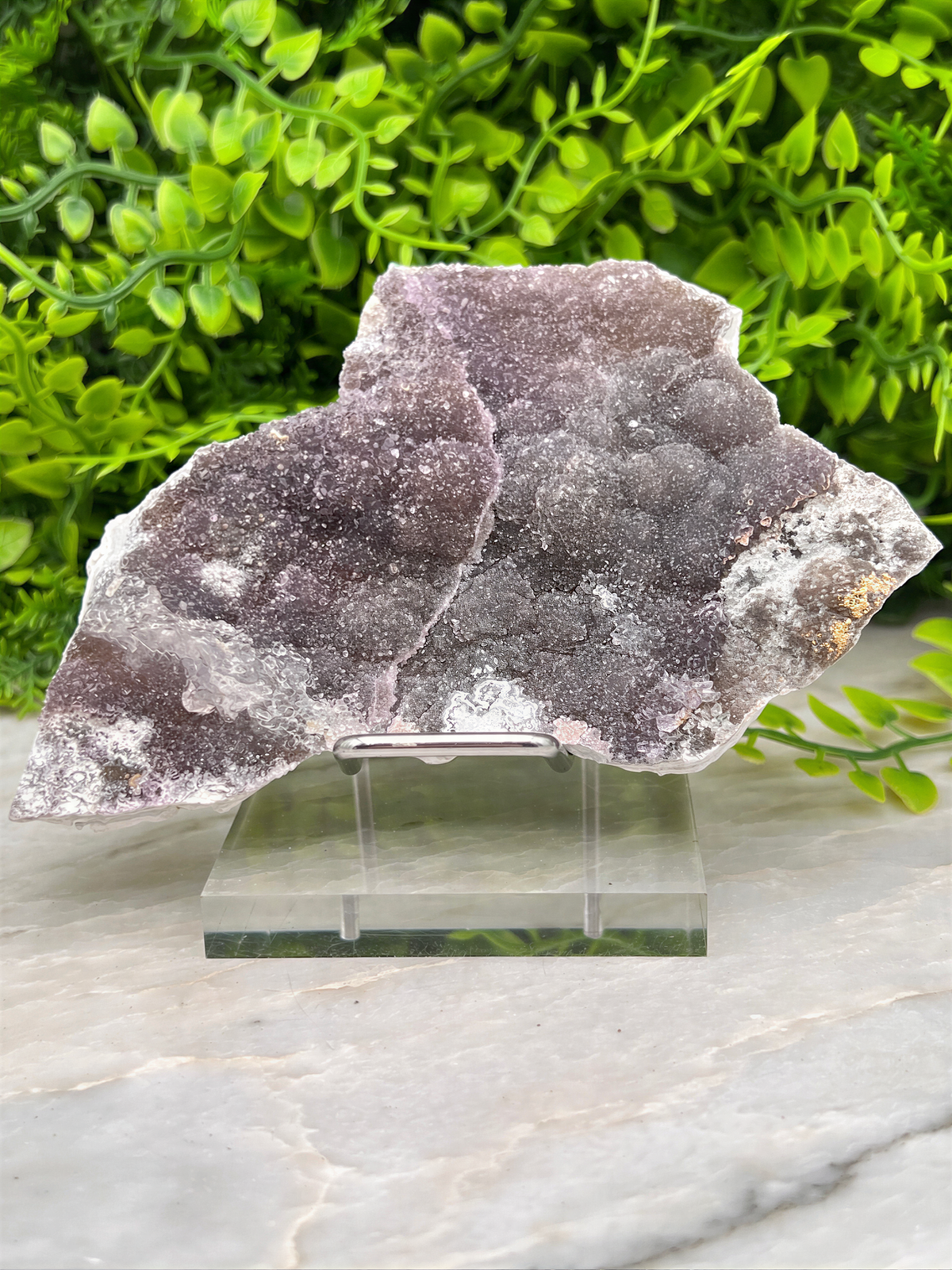 a piece of amethyst, a purple crystal, displayed on a clear glass stand. The crystal is surrounded by greenery, creating a natural and serene setting.