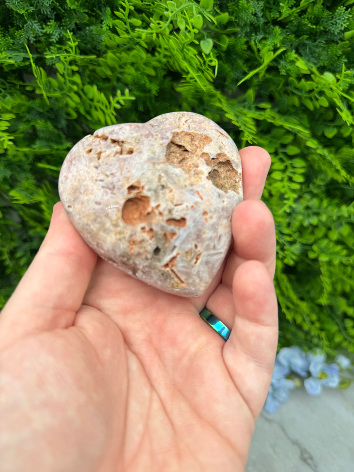 A hand holding a heart-shaped rock against a backdrop of green foliage.