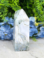 A jadeite crystal with a unique, textured surface sits in the center of the image, surrounded by vibrant blue flowers.