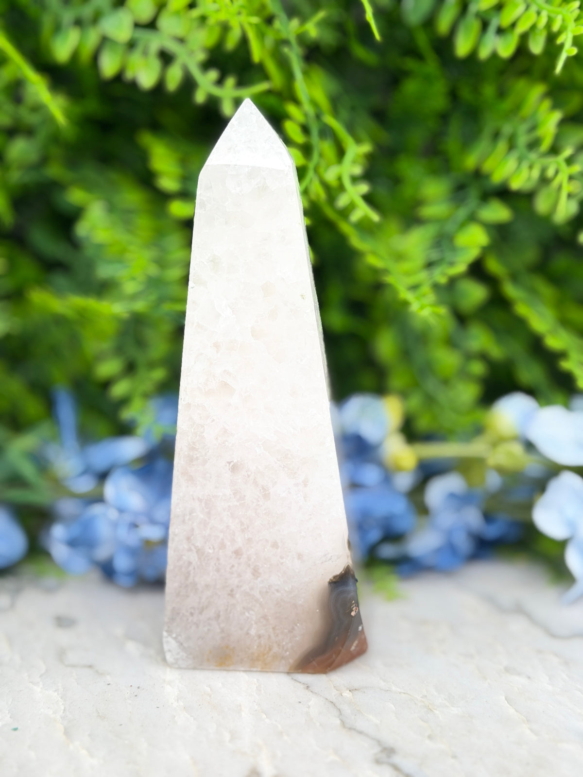 A white pyramid-shaped crystal sits on a white surface, surrounded by lush green foliage and blue flowers.