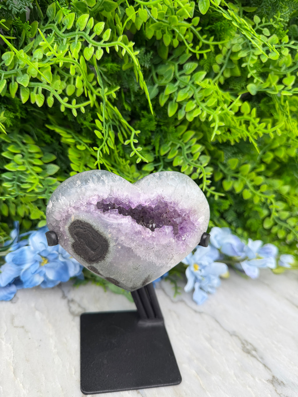 A heart-shaped amethyst geode is displayed on a black stand, surrounded by lush green foliage and blue flowers.