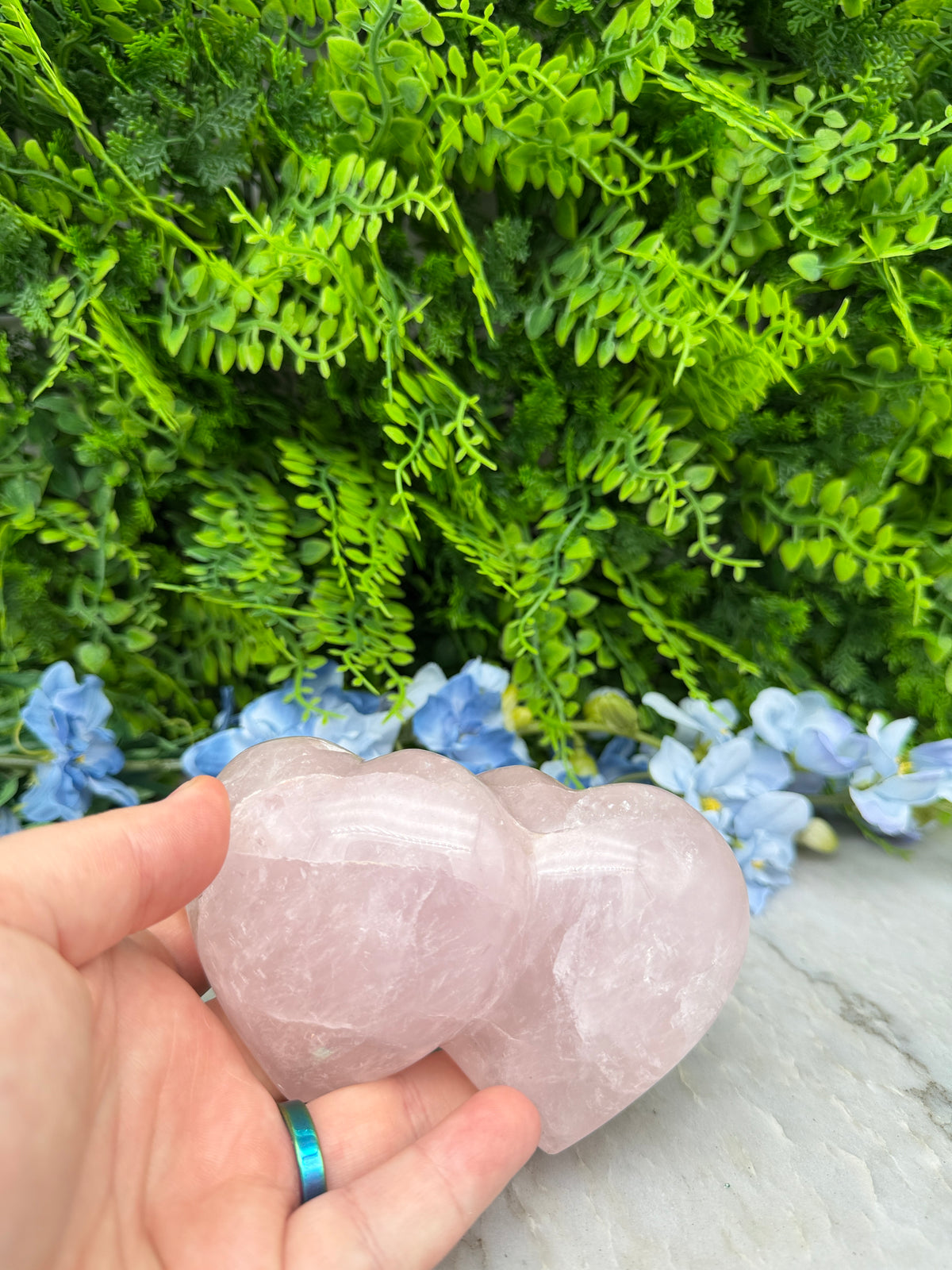 A hand holding a pink crystal next to a green plant with blue flowers.