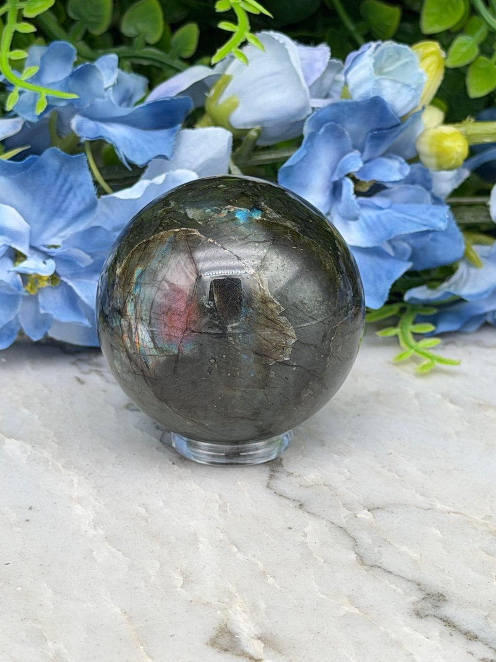 a close-up view of a dark, textured stone sphere resting on a white surface, surrounded by vibrant blue flowers.