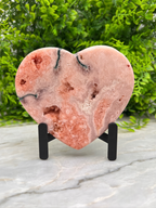 A pink heart-shaped rock with a unique, textured surface sits on a marble surface, surrounded by lush green foliage.