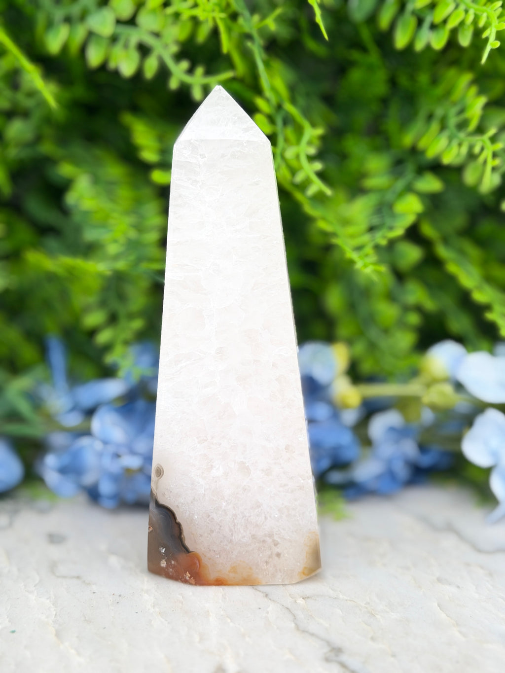 A white pyramid-shaped crystal sits on a white surface, surrounded by lush green foliage.