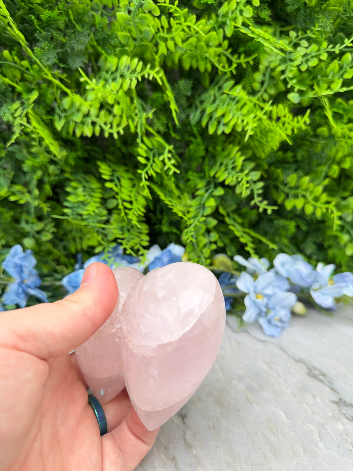 A hand holding a pink crystal against a backdrop of green foliage and blue flowers.