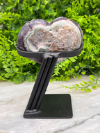 A heart-shaped amethyst geode on a black metal stand, placed on a marble surface with a green plant in the background.