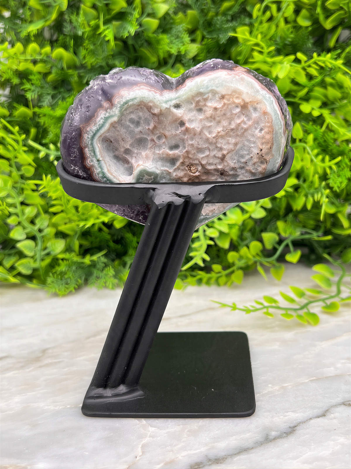 A heart-shaped amethyst geode on a black metal stand, placed on a marble surface with a green plant in the background.