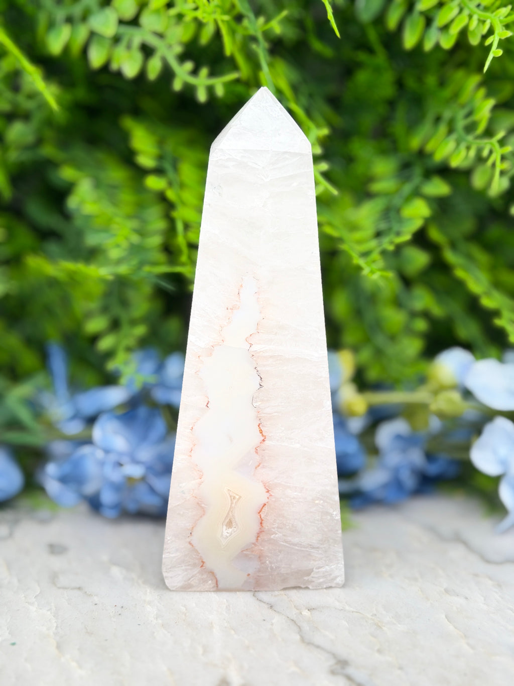 A white, jagged-edged crystal stands in the center of a lush, green background, surrounded by blue flowers.