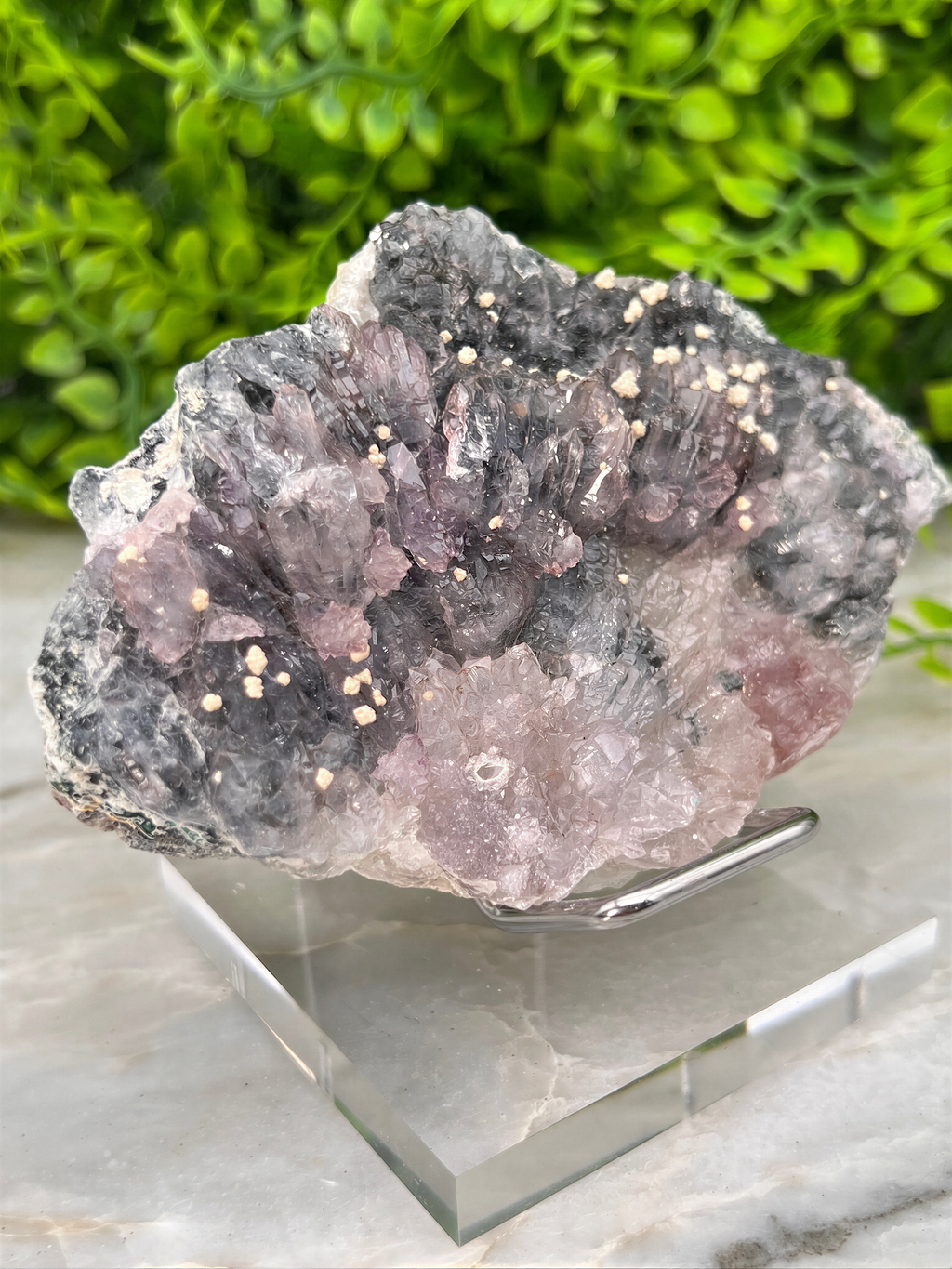 A clear glass display case containing a large, irregularly shaped crystal with a mix of gray, pink, and white colors. The crystal appears to be a type of quartz or feldspar, with a rough, natural texture. The display case is placed on a marble surface, and some greenery is visible in the background.